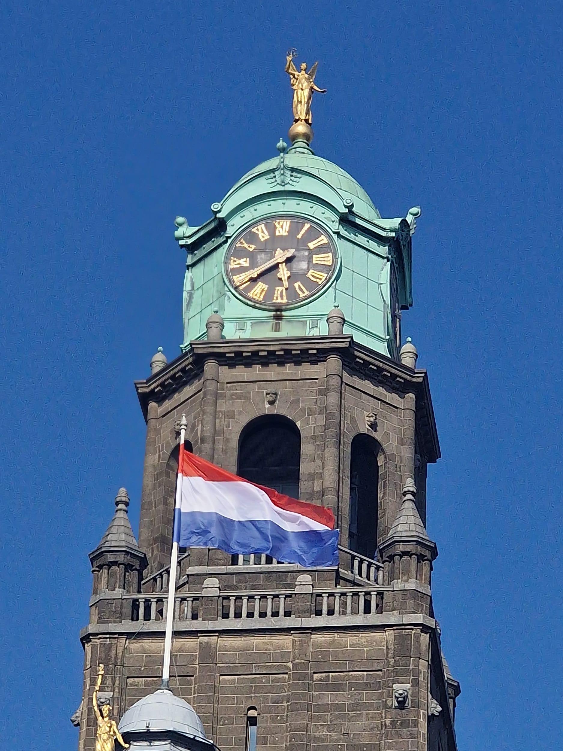 A church tower with a green copper dome and gilded clock face, a golden figure on its spire and the Dutch flag flying below, against a cloudless blue sky