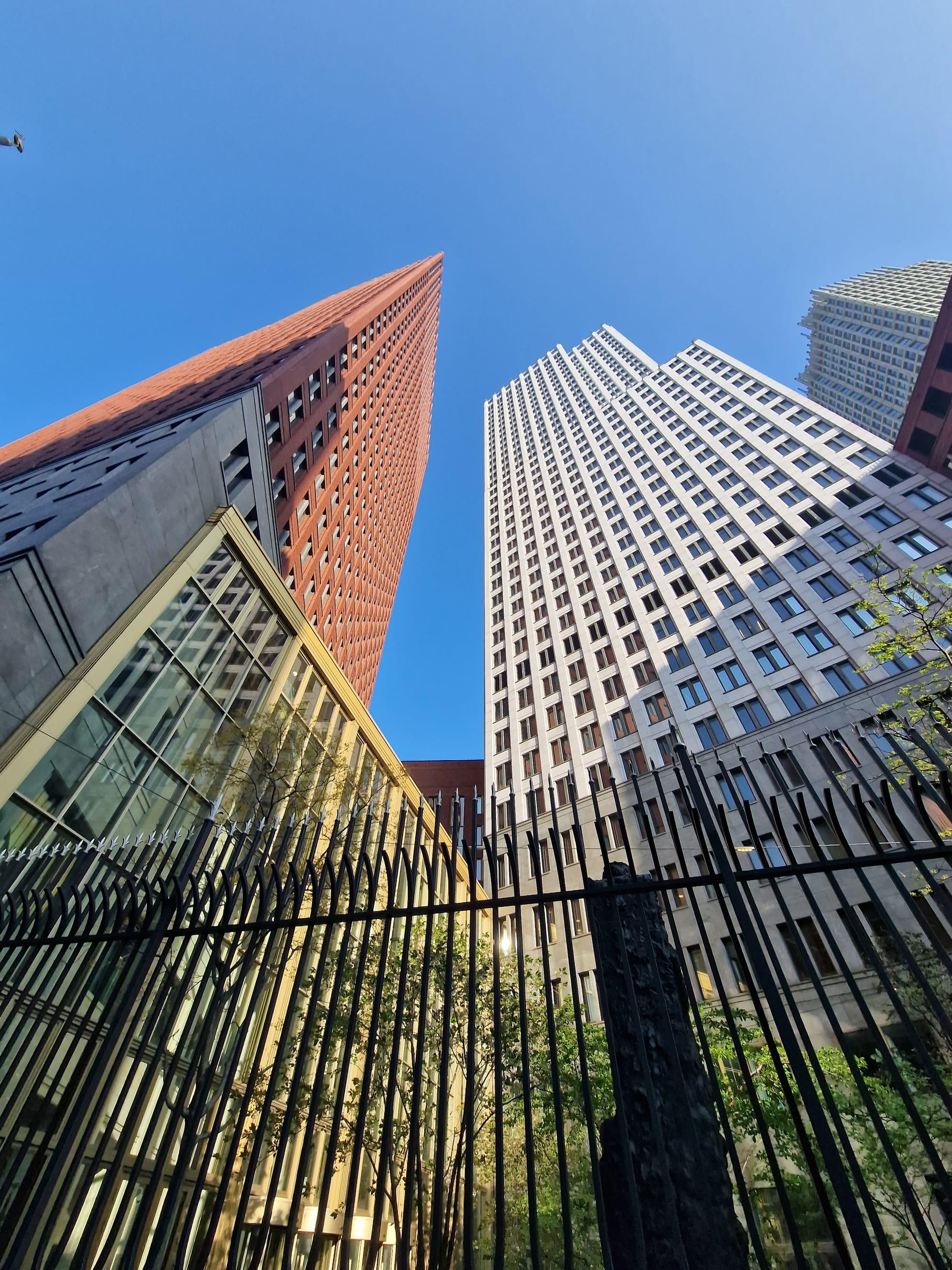 The same pair of towers framed from behind tall black railings, a glazed roof catching the light in the foreground