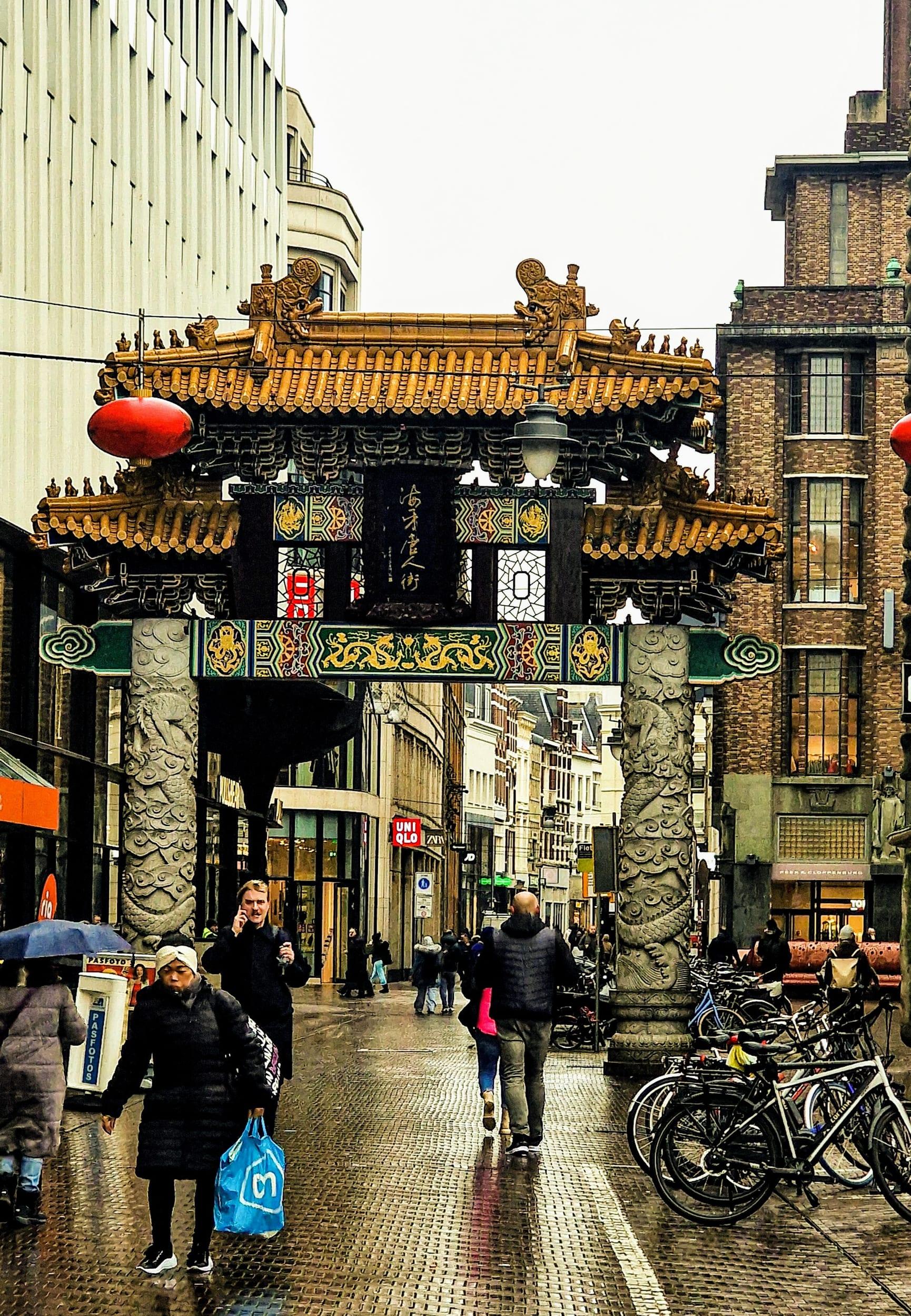 An ornate Chinese gate spanning a shopping street on a wet afternoon, people walking beneath it under umbrellas and bicycles parked along the kerb