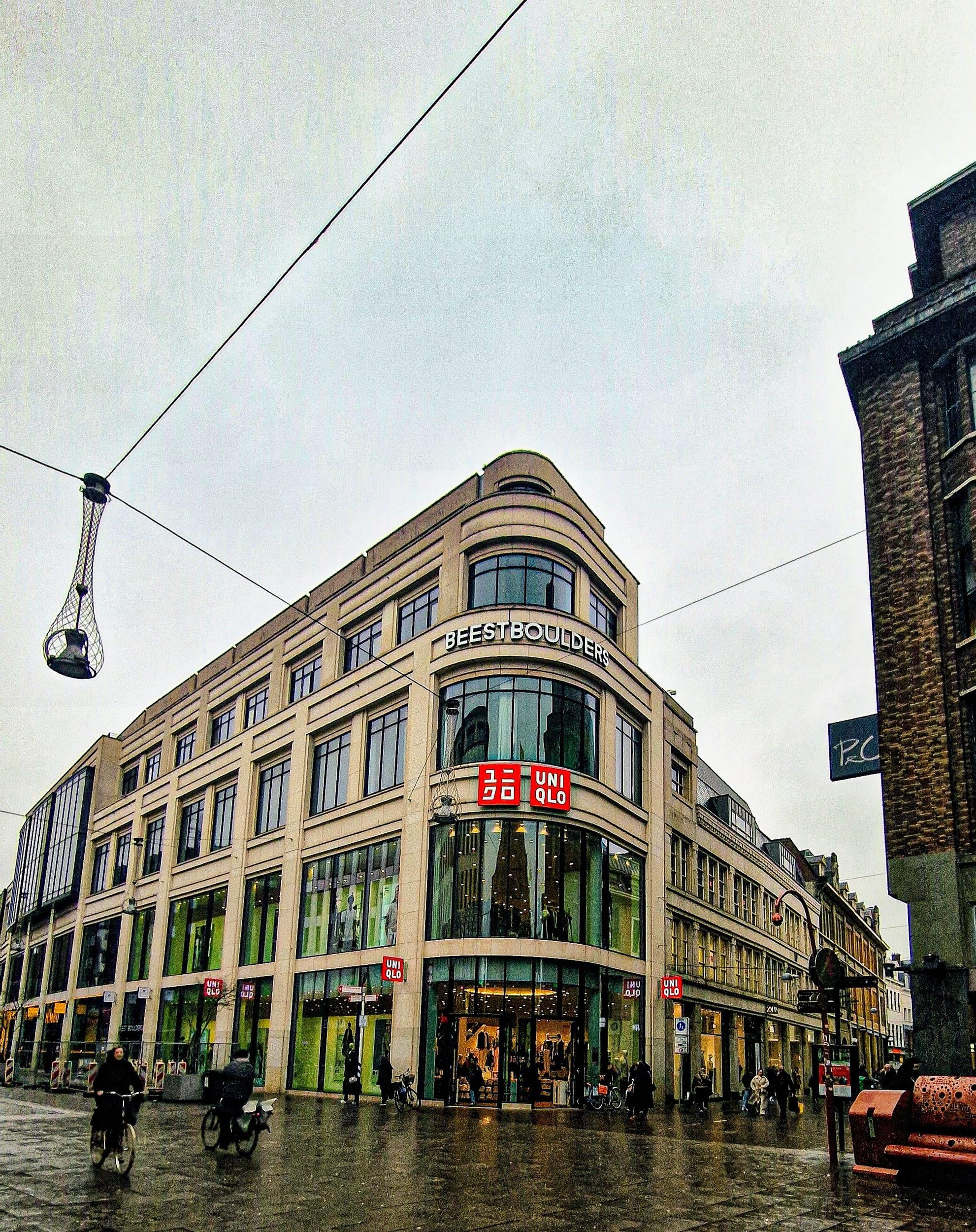 A curved corner building on a rain-slicked street, cyclists crossing in front of its lit shopfronts under a flat grey sky