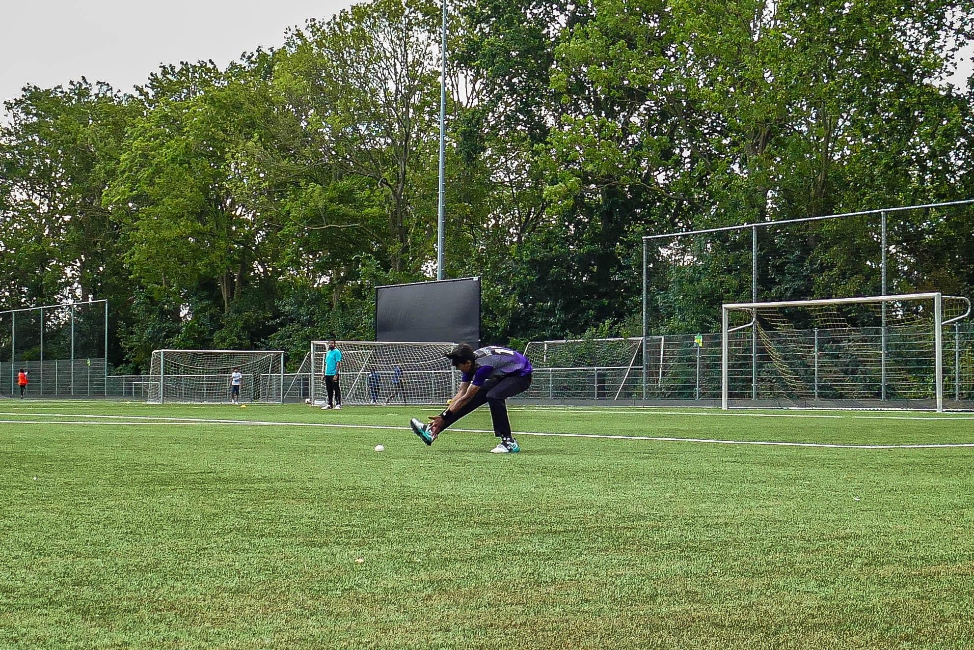 A player bending to the ball on an empty green pitch, goalposts and a line of trees behind