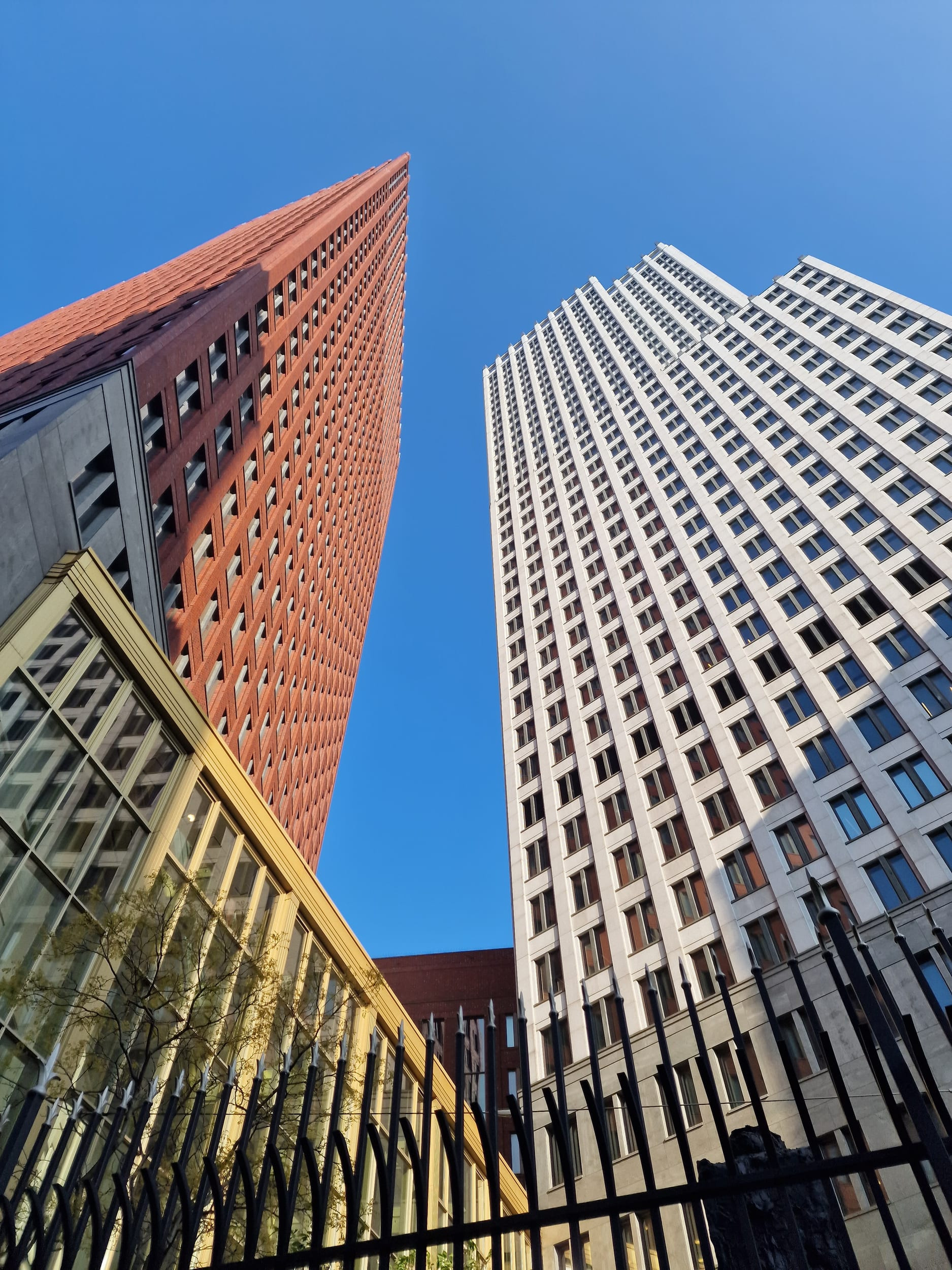 Two high-rise towers seen from street level, one clad in red brick and one in white, converging towards a clear blue sky